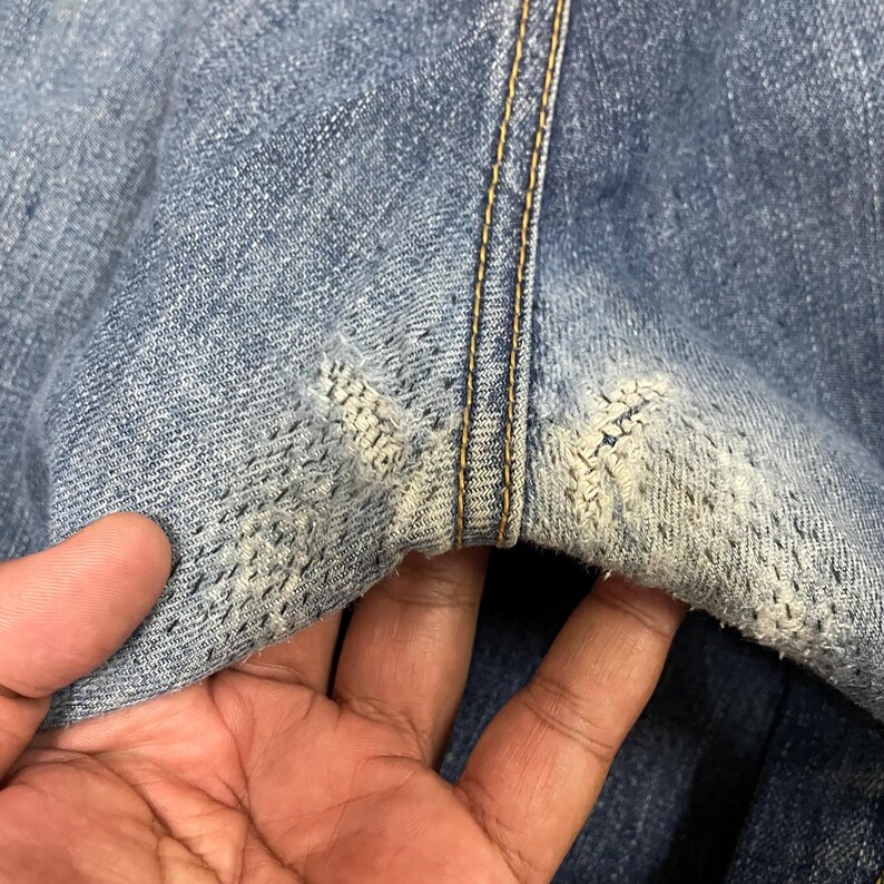 May include: A pair of blue denim jeans featuring a prominent vertical seam with gold-colored stitching. The fabric displays two distinct areas of wear and fraying near the seam, revealing lighter blue threads and a distressed texture.