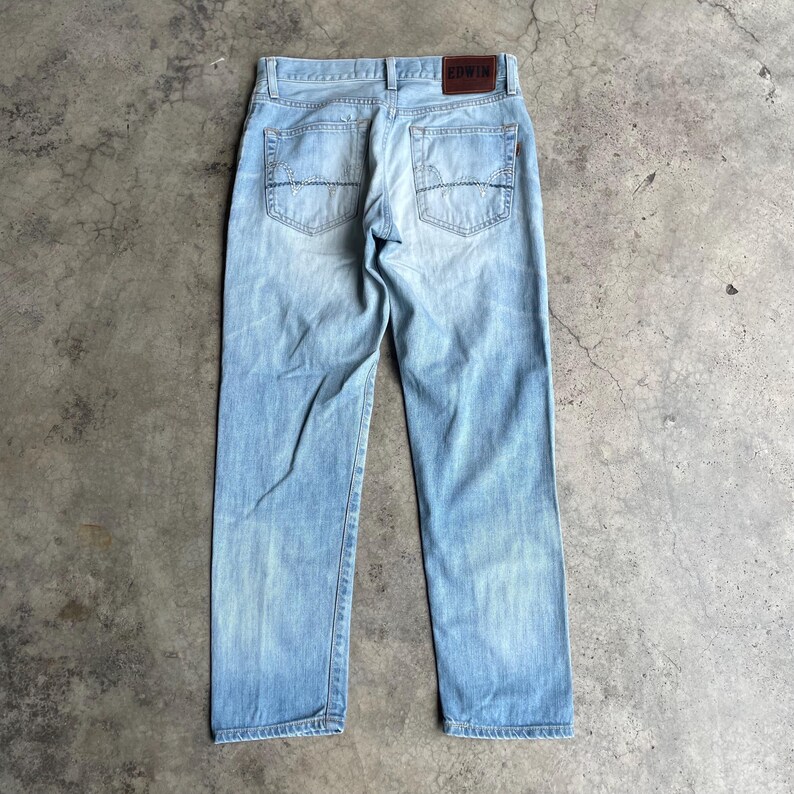 May include: Pale blue denim jeans with a faded effect. The jeans have a classic five-pocket design, a brown leather patch with the brand name, and a straight leg fit. The back pockets feature decorative stitching.