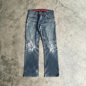 K&ouml;nnte beinhalten: Verblasste blaue Jeans mit abgenutzten Details, mit zerrissenen Bereichen an beiden Knien. Die Jeans haben ein klassisches F&uuml;nf-Taschen-Design, einen Knopf- und Rei&szlig;verschluss und einen roten Bund. Die Jeans liegen flach auf einer Betonoberfl&auml;che.