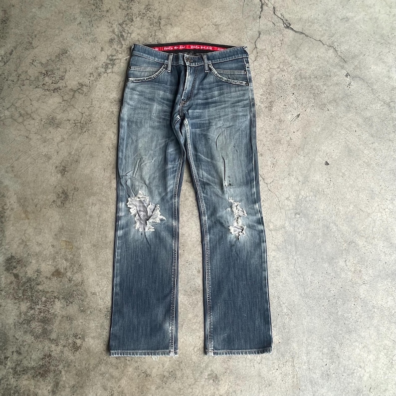 K&ouml;nnte beinhalten: Verblasste blaue Jeans mit abgenutzten Details, mit zerrissenen Bereichen an beiden Knien. Die Jeans haben ein klassisches F&uuml;nf-Taschen-Design, einen Knopf- und Rei&szlig;verschluss und einen roten Bund. Die Jeans liegen flach auf einer Betonoberfl&auml;che.