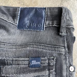 May include: Close-up of a pair of gray denim jeans. The jeans feature a dark blue leather patch at the waistband with the text "JERSEYS". A smaller patch on the back pocket reads "EDWIN JERSEYS".