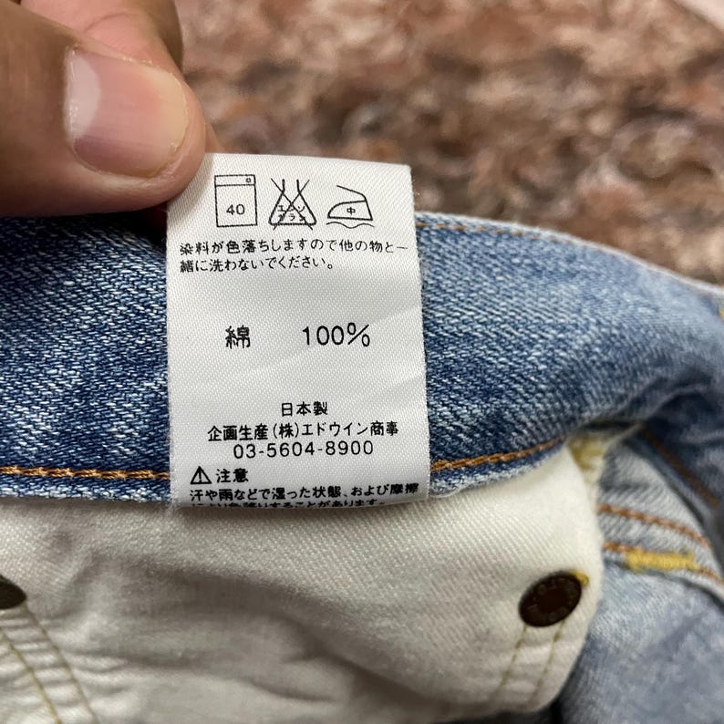 May include: Close-up of a tag on denim fabric. The tag displays washing instructions, including a 40-degree wash symbol. Text on the tag indicates the fabric is 100% cotton and provides Japanese manufacturing details.