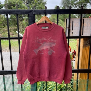 May include: A maroon sweatshirt with the text "Anglers Guide" and a graphic of a fish and fishing rod. The sweatshirt is hanging on a wooden hanger against a black metal fence.