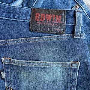 Op de afbeelding: Close-up van een blauwe spijkerbroek met een achterzak en een leren patch. De patch is donkerbruin met de merknaam "EDWIN" in rode letters en de tekst "MADE IN JAPAN".