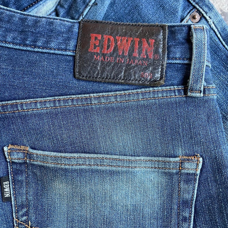 Op de afbeelding: Close-up van een blauwe spijkerbroek met een achterzak en een leren patch. De patch is donkerbruin met de merknaam "EDWIN" in rode letters en de tekst "MADE IN JAPAN".