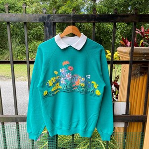May include: Teal sweatshirt with a white collar and floral design. The sweatshirt features a colorful array of flowers in shades of yellow, pink, orange, and purple. The flowers are arranged in a horizontal pattern across the chest.