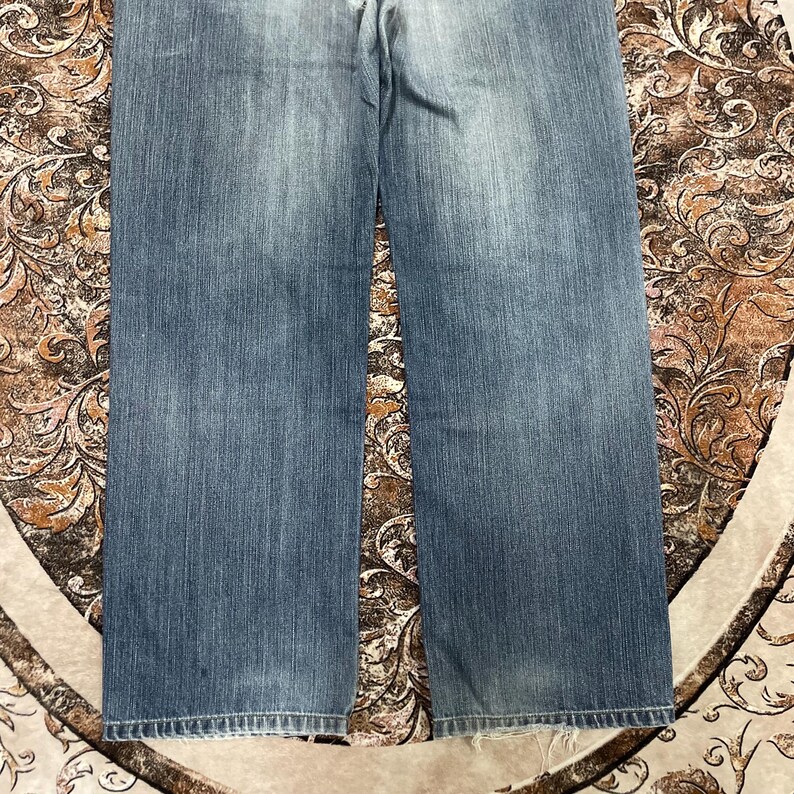 May include: A pair of blue denim jeans featuring a faded wash effect across the thighs and straight-cut legs. The fabric displays a classic twill texture with visible stitching along the bottom hems, which show slight signs of wear and fraying.