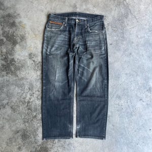 May include: Dark wash denim jeans with a straight leg cut. The jeans feature a classic five-pocket design, a button and zip closure, and a small brown leather patch detail on the front pocket. The jeans are distressed with faded areas.