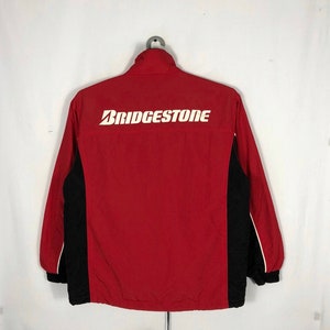 Może przedstawiać: Czerwono-czarna kurtka wiatrówka z białym logo "BRIDGESTONE" z tyłu.