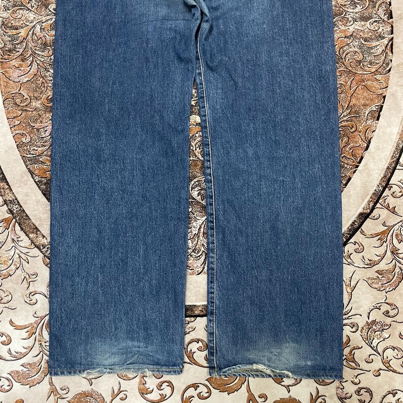 May include: A pair of blue denim jeans featuring a classic straight-leg cut with visible fading on the lower legs and frayed, worn edges at the bottom hems. The fabric displays a medium blue wash with traditional orange-toned stitching details.