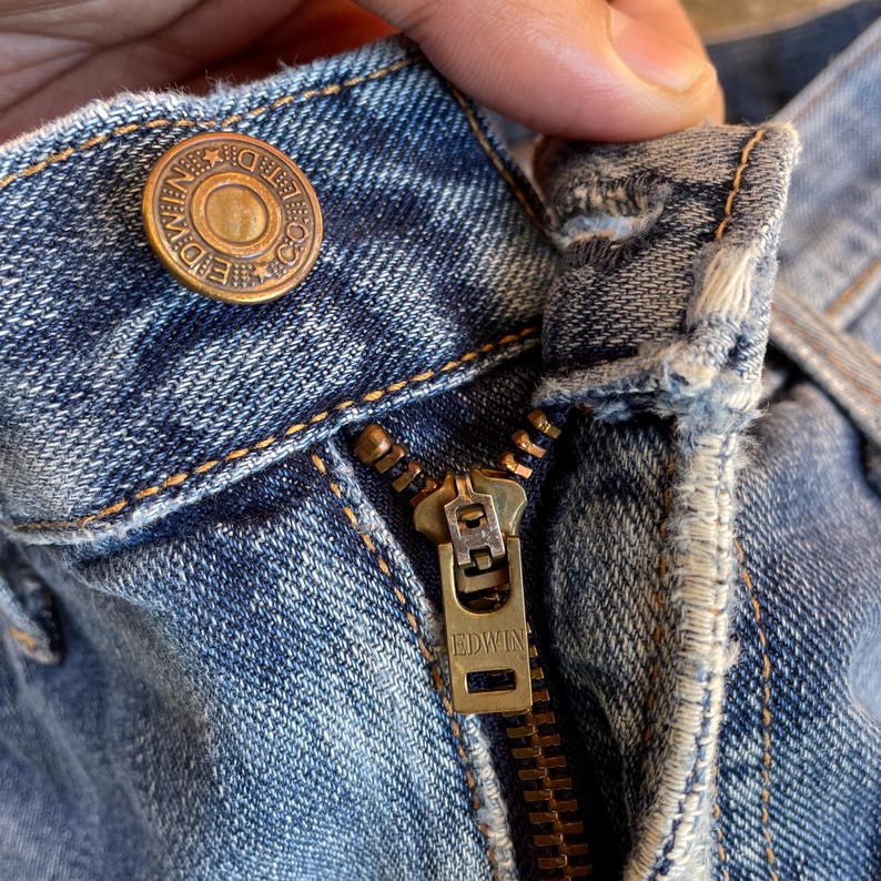 Pu&ograve; includere: Primo piano di jeans blu con cerniera e bottone in ottone. Il bottone &egrave; impresso con il marchio "EDWIN". La cerniera &egrave; anch'essa in ottone e riporta il marchio "EDWIN". I jeans sono usurati e sbiaditi.