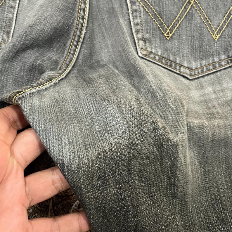 May include: Close-up of gray denim jeans with a faded appearance. The jeans have a classic five-pocket design, with a back pocket featuring a signature stitching pattern. The stitching is in a contrasting yellow color.