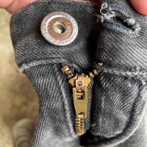 May include: Close-up of a pair of gray denim jeans with a brass zipper and button. The button is silver with the text "EDWIN COMPANY EST. 1961". The zipper is partially open, revealing the teeth.