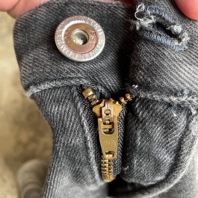 May include: Close-up of a pair of gray denim jeans with a brass zipper and button. The button is silver with the text "EDWIN COMPANY EST. 1961". The zipper is partially open, revealing the teeth.