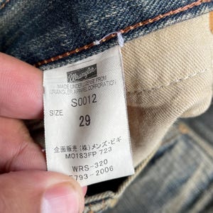 May include: Close-up of a Wrangler jeans tag. The tag is white with black text, including the brand name, size 29, and other details. The jeans are a faded blue denim.