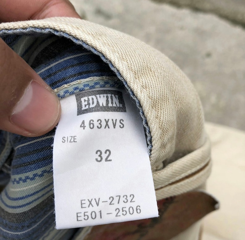 May include: A white and blue striped fabric label with the text "EDWIN" in black, "463XVS" in black, "SIZE" in black, "32" in black, "EXV-2732" in black, and "E501-2506" in black.