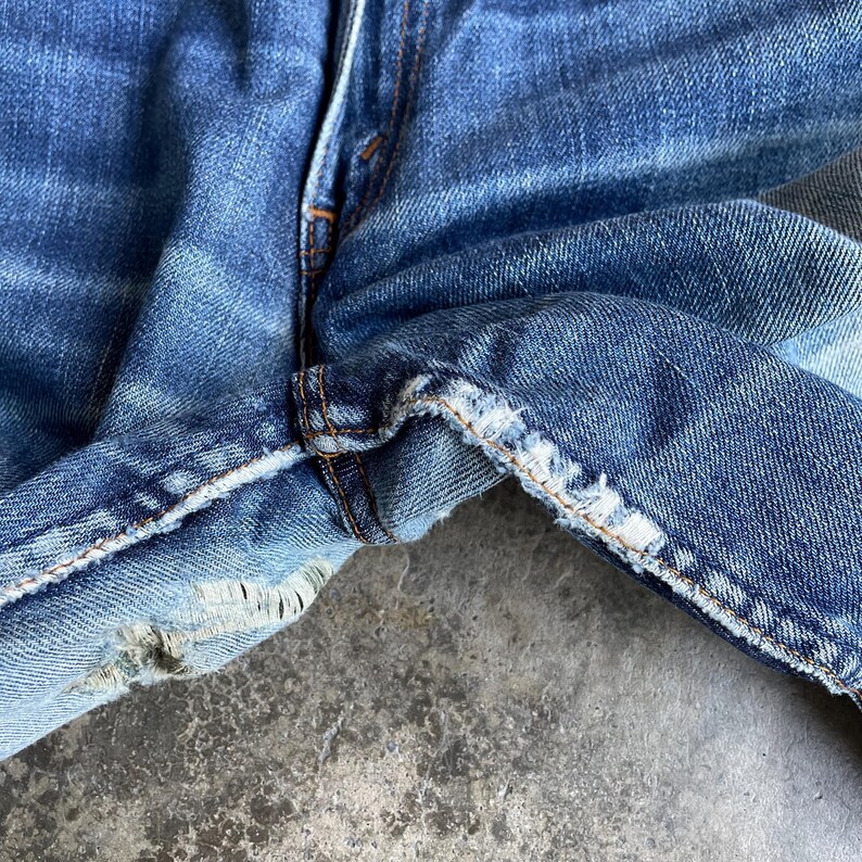 May include: Close-up of worn blue denim jeans. The image highlights the distressed details, including frayed edges and visible stitching. The fabric shows signs of wear and tear, with faded areas and a vintage aesthetic.