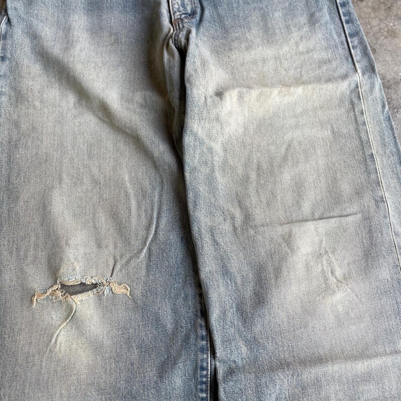 May include: Light blue denim jeans with a distressed, faded appearance. The jeans have a ripped knee, showing the frayed edges of the fabric. The denim has a worn look, with lighter patches and subtle variations in color.