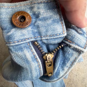 May include: Close-up of light blue denim jeans with a brass zipper and a button closure. The button is brown with the word "EDWIN" embossed on it. The zipper is partially open, revealing the dark blue fabric underneath.