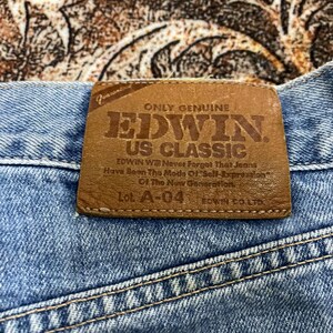 May include: A pair of light blue denim jeans featuring a brown leather patch on the back waistband. The patch is embossed with the text: ONLY GENUINE EDWIN US CLASSIC, EDWIN Will Never Forget That Jeans Have Been The Mode Of Self-Expression Of The New Generation, Lot. A-04, EDWIN CO. LTD.