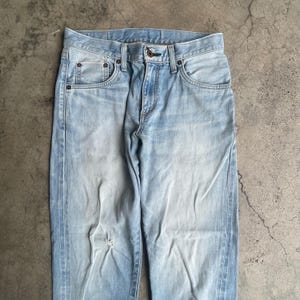 May include: Light blue denim jeans with a faded wash and a distressed knee. The jeans feature classic five-pocket styling, a button and zip closure, and belt loops. The denim has a worn appearance.