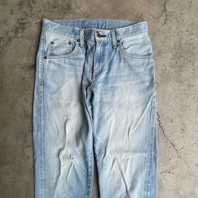May include: Light blue denim jeans with a faded wash and a distressed knee. The jeans feature classic five-pocket styling, a button and zip closure, and belt loops. The denim has a worn appearance.