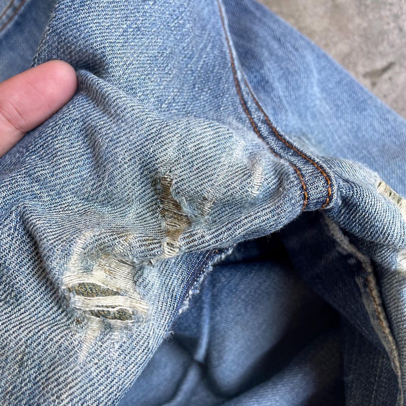 May include: Close-up of distressed light blue denim jeans. The fabric shows wear and tear with visible rips and frayed edges. Brown stitching is visible along the seams, highlighting the construction of the garment.
