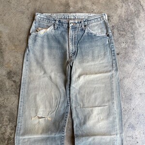 May include: Faded light blue denim jeans with a distressed look. The jeans have a relaxed fit, a button and zipper closure, and classic five-pocket styling. There is a tear on the left knee and a worn area on the left pocket.