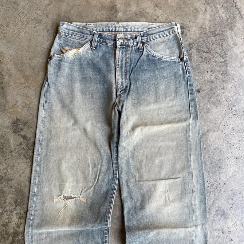 May include: Faded light blue denim jeans with a distressed look. The jeans have a relaxed fit, a button and zipper closure, and classic five-pocket styling. There is a tear on the left knee and a worn area on the left pocket.