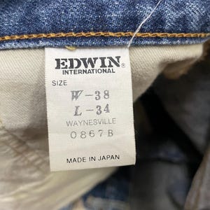 May include: A white interior garment tag on blue denim fabric with the text EDWIN INTERNATIONAL, SIZE W-38, L-34, WAYNESVILLE, 0867B, and MADE IN JAPAN. The tag is stitched into the waistband with orange thread, showing clear black printed text.