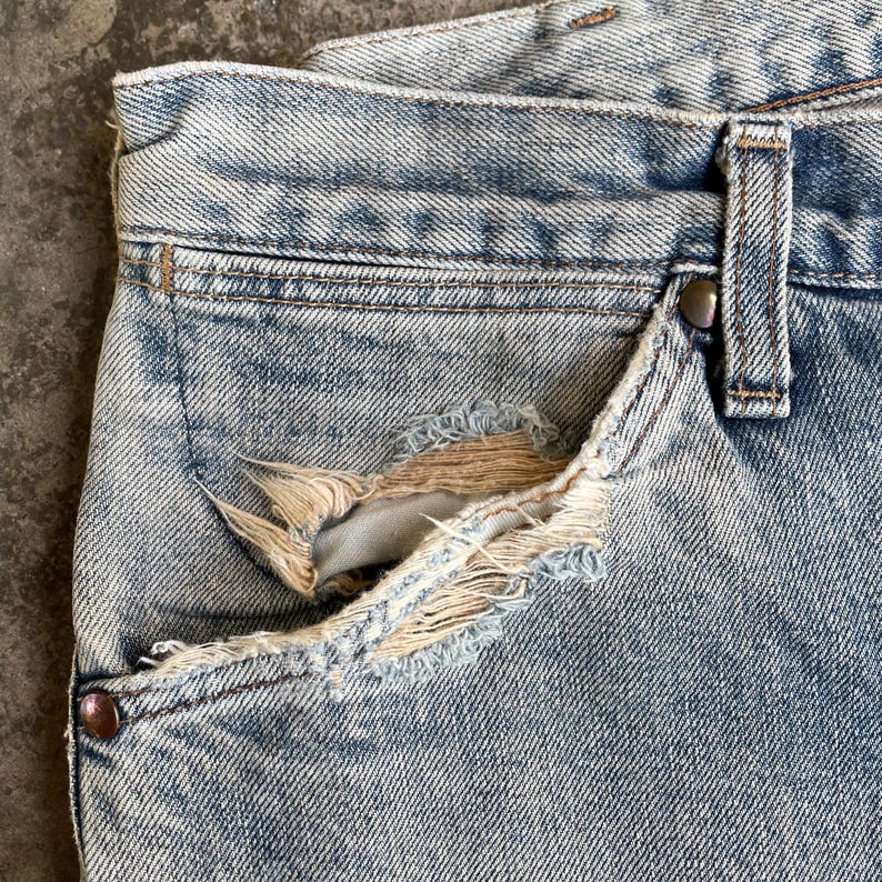 May include: Close-up of light blue denim jeans with a distressed pocket. The pocket has a torn section revealing the interior. The jeans show signs of wear and fading, with a metal button and belt loop visible.