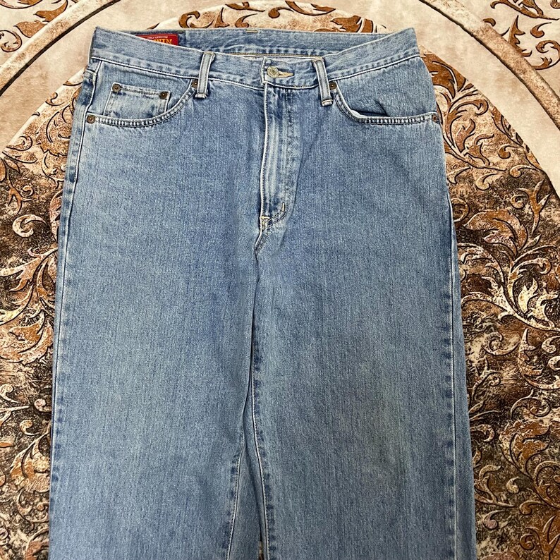 May include: Light blue denim jeans featuring a classic five-pocket design, a metal button closure, a zippered fly, and a small red rectangular label with the text EDWIN stitched onto the interior waistband near the back center.