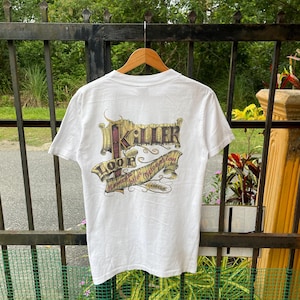 Op de afbeelding: Wit t-shirt met een grafisch ontwerp op de achterkant. Het ontwerp toont het woord "Killer" in een gestileerd lettertype boven het woord "Loop" en een banner met de tekst "before restoration of love".