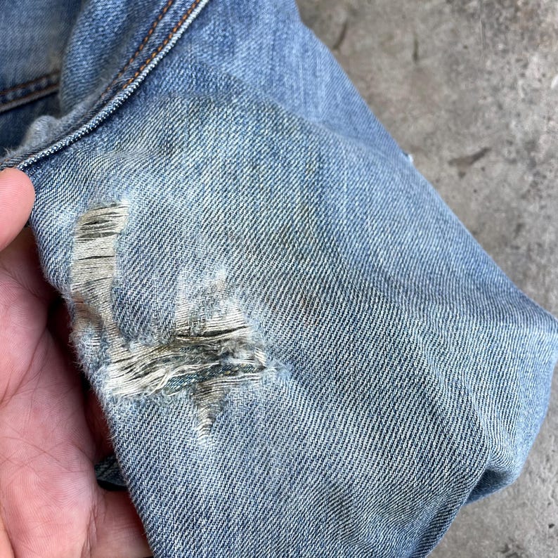 May include: Close-up of light blue denim fabric with a distressed, ripped detail. The fabric shows signs of wear and tear, with visible threads and a faded appearance. The image highlights the texture and colour variations of the denim.