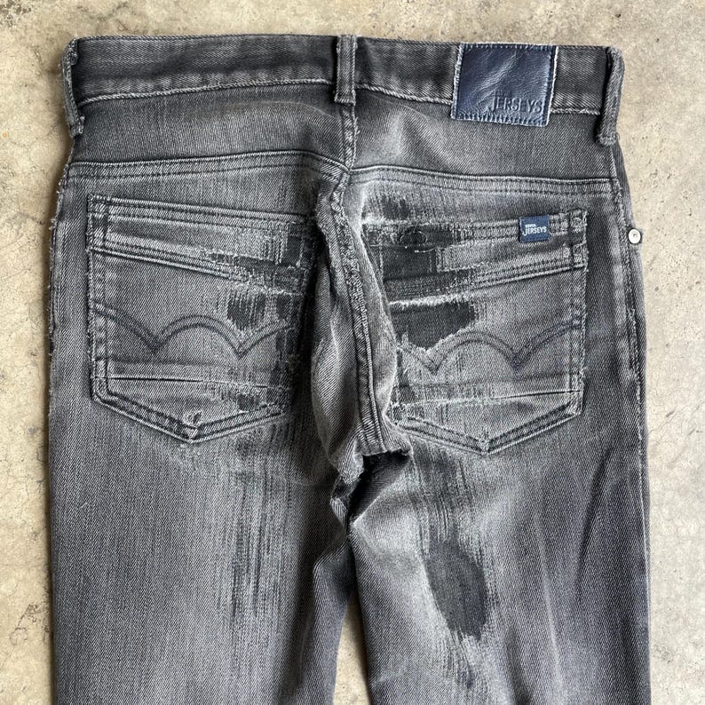 May include: Close-up of distressed black denim jeans. The jeans feature a faded wash, visible wear and tear, and the word "JERSEYS" on a leather patch. The back pockets have a distinctive design.