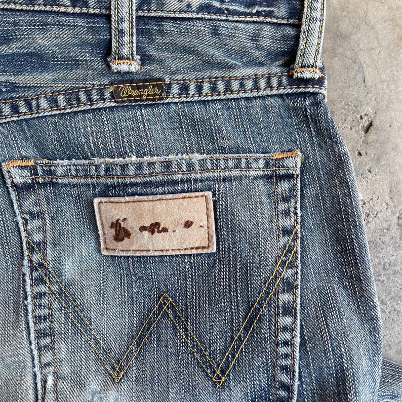 May include: Close-up of blue denim jeans with the Wrangler logo on the back pocket. The jeans have a worn, distressed look with yellow stitching and a leather patch. The back pocket features the iconic Wrangler "W" stitching.