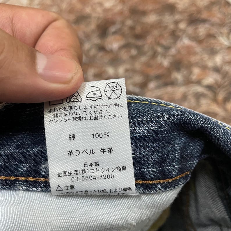 May include: A white rectangular garment care label sewn into dark blue denim fabric. The label features black Japanese text, care symbols for washing and ironing, and specifies 100% cotton composition with a cowhide leather label component.