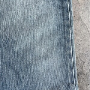 May include: Close-up of light blue denim fabric. The denim has a faded appearance with subtle variations in colour and texture. The image shows a portion of the jeans, including a seam.