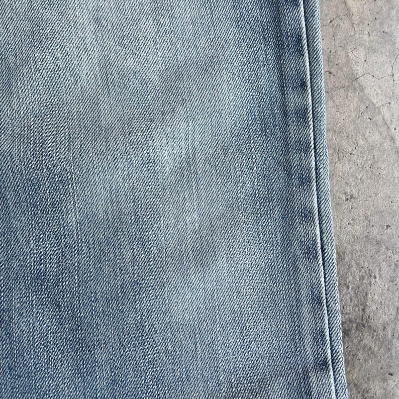 May include: Close-up of light blue denim fabric. The denim has a faded appearance with subtle variations in colour and texture. The image shows a portion of the jeans, including a seam.