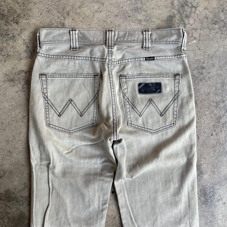 K&ouml;nnte beinhalten: Nahaufnahme der R&uuml;ckseite einer hellgrauen Wrangler-Jeans. Die Jeans hat schwarze N&auml;hte, zwei Ges&auml;&szlig;taschen mit dem Wrangler "W"-Logo und einen schwarzen Aufn&auml;her mit dem Wrangler-Logo. Die Jeans ist aus strapazierf&auml;higem Denim-Material.