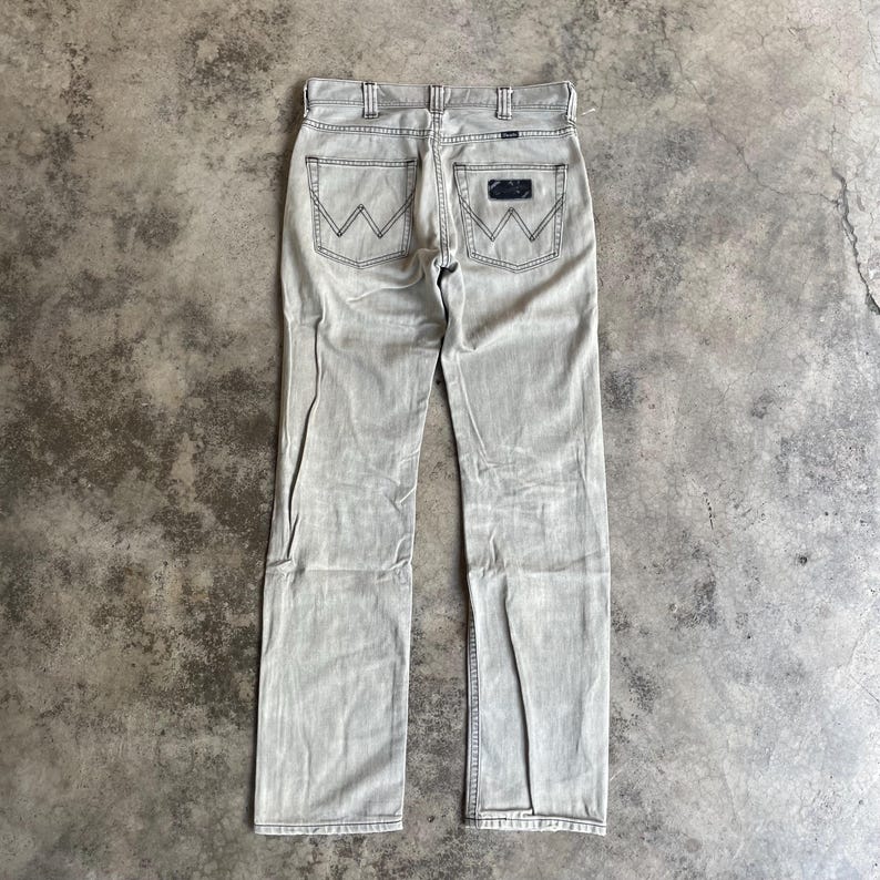 K&ouml;nnte beinhalten: Hellgraue Jeans mit dem Wrangler-Logo auf den Ges&auml;&szlig;taschen. Die Jeans haben ein klassisches F&uuml;nf-Taschen-Design und eine gerade Beinpassform. Die Ges&auml;&szlig;taschen haben die ikonischen "W" -N&auml;hte.