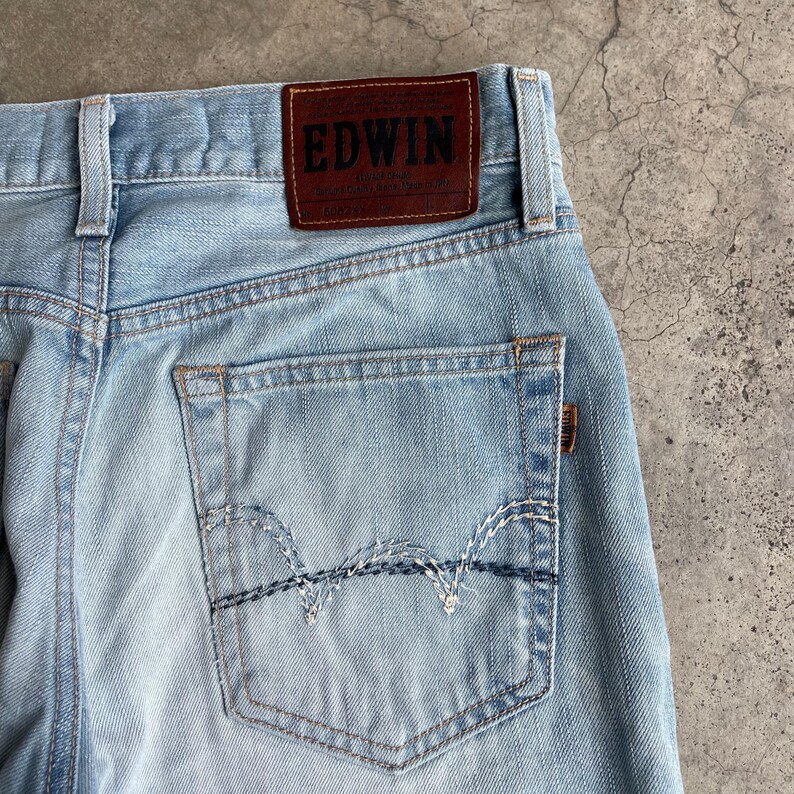 May include: Pale blue denim jeans featuring a brown leather "EDWIN" patch. The jeans have a classic five-pocket design with a decorative stitch on the back pocket. The denim has a faded appearance.
