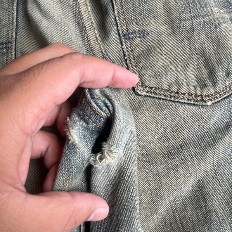 May include: Close-up of a distressed denim jacket. The fabric is faded blue with visible wear and tear, including a hole near the seam. The pocket and stitching are visible.