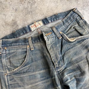May include: Close-up of a pair of faded blue denim jeans. The jeans have a classic five-pocket design, with visible wear and tear, and a distressed look. The brand name "BEN" is visible on a label.