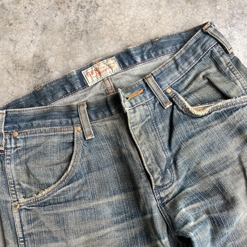 May include: Close-up of a pair of faded blue denim jeans. The jeans have a classic five-pocket design, with visible wear and tear, and a distressed look. The brand name "BEN" is visible on a label.
