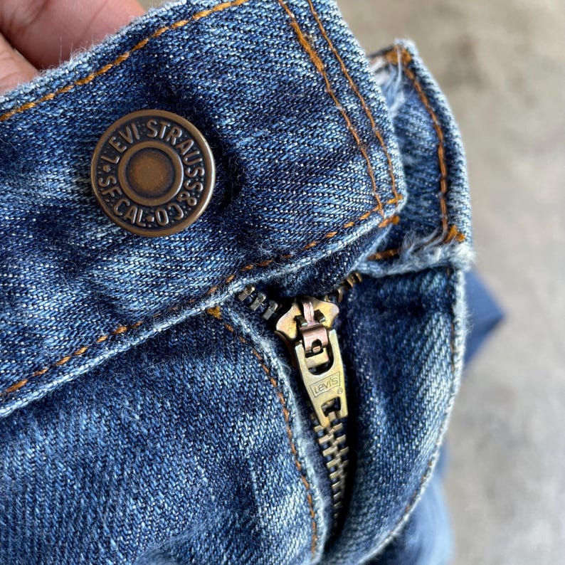 May include: Close-up of blue denim jeans with a brass button and zipper. The button is embossed with "LEVI STRAUSS & CO. CAL. 03". The zipper is metallic with the Levi's logo.