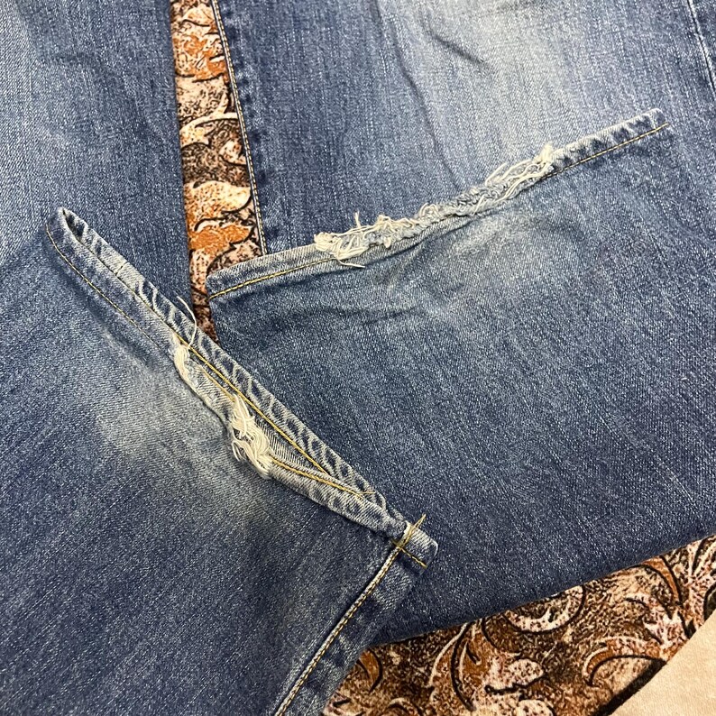 May include: A pair of blue denim jeans featuring a medium wash with visible fading and distressed, frayed hems at the bottom of the pant legs. The fabric shows a classic twill texture with gold-toned stitching along the seams and leg openings.