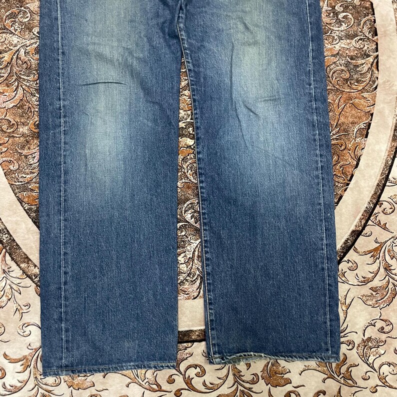 May include: A pair of blue denim jeans featuring a classic straight-leg cut with a medium wash finish. The fabric displays subtle fading on the front panels, visible vertical stitching along the outer seams, and a standard hem at the bottom edges.