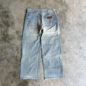 May include: Light blue denim Wrangler jeans with a faded wash. The jeans have a classic five-pocket design, a brown leather patch, and a straight leg. The jeans are worn and have a distressed look.
