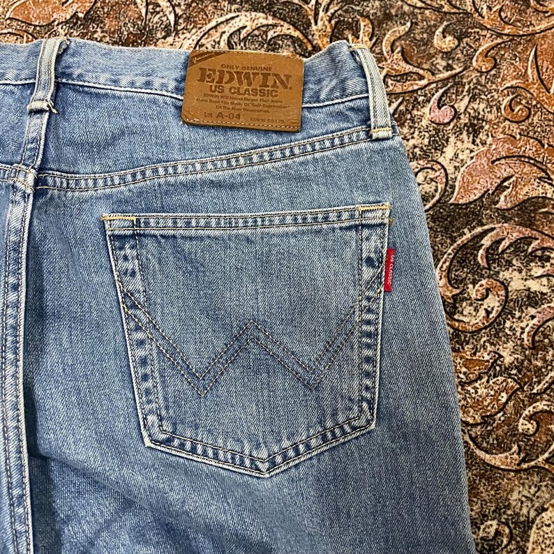 May include: Light blue denim jeans featuring a tan leather patch on the waistband embossed with the text EDWIN US CLASSIC. The back pocket displays a signature W-shaped stitch pattern and a small red tab with the text US CLASSIC.